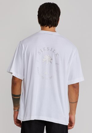 Mann trägt ein weißes, übergroßes T-Shirt mit einem bestickten Palmenmotiv und dem Schriftzug "SIKSILK" sowie "FOLLOW THE MOVEMENT" auf der Rückseite.