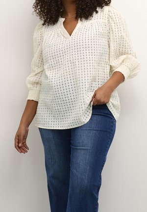Vrouw met krullend haar draagt een witte geruite blouse met pofmouwen en blauwe spijkerbroek, staand tegen een effen lichte achtergrond.