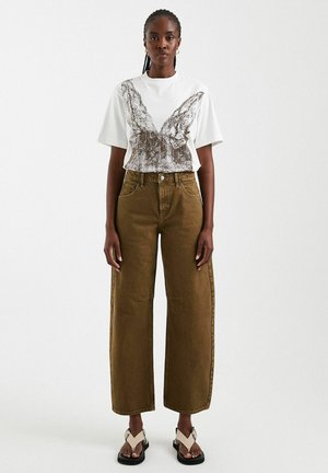 Woman with braided hair wearing a white graphic t-shirt, high-waisted wide-leg brown pants, and two-tone open-toe sandals standing against white background.