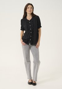 Schwarze Bluse mit Rüschendetails, kurzen Ärmeln und Knopfschluss vorne, kombiniert mit hellgrauen, geraden Jeans und schwarzen Schuhen.