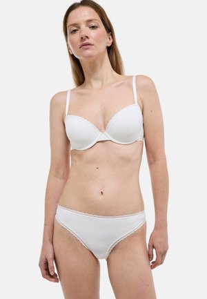 Femme à la peau claire et aux cheveux bruns portant un soutien-gorge blanc rembourré et une culotte blanche assortie bordée de dentelle sur un fond uni.