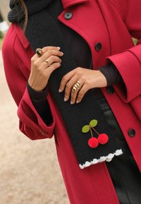 Černý žebrovaný šátek s bílým lemem, zdobený červenými plsťovými třešněmi a zelenými listy, nošený s jasně červeným kabátem a několika zlatými prsteny.