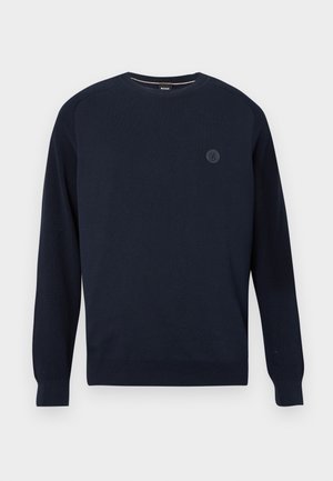 Pull à col rond bleu marine en tissu tricoté, avec des poignets et un ourlet côtelés. Un logo circulaire est positionné sur la poitrine.