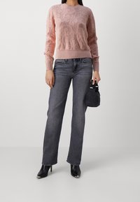 Pull rose tricoté avec motif floral, poignets côtelés, assorti à un jean gris à jambes droites et des bottines à talons noires, tenant un petit sac noir.