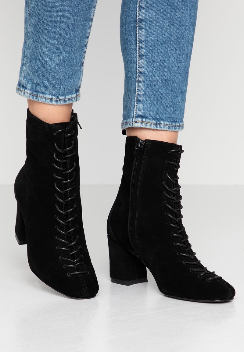 Bottes cheville en daim noir avec talons carrés, lacets à l'avant, fermeture éclair sur le côté, portées avec un jean bleu coupé au-dessus de la cheville sur une surface claire.