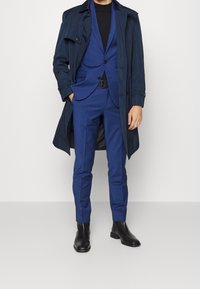 Marineblauwe trenchcoat over een blauw pak met een getailleerd ontwerp, zwarte coltrui en zwarte laarzen. Eenvoudig, gladde textuur zonder zichtbare patronen.