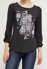 Žena v černém dlouhém rukávu s bílými květinovými vyšívanými vzory a tmavě modrými džínami, stojící proti jednobarevnému pozadí.