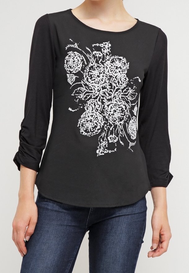 Žena v černém dlouhém rukávu s bílými květinovými vyšívanými vzory a tmavě modrými džínami, stojící proti jednobarevnému pozadí.