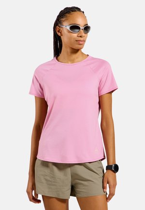 Persona con occhiali da sole argentati, camicia rosa a maniche corte, pantaloncini beige e uno smartwatch nero, in piedi contro uno sfondo bianco semplice.