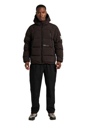 Schwarze isolierte Steppjacke mit Kapuze, ausgestattet mit Reißverschlusstaschen und einem Logo auf der Brust. Getragen mit schwarzen Hosen und klobigen Turnschuhen.