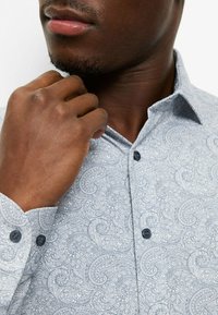 Hellblaues Hemd mit Knopfleiste, Paisley-Muster, strukturierter Stoff und dunkle Knöpfe an Bündchen und Kragen.