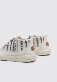 Zapatillas blancas informales con una parte superior de tela estampada, que presenta diseños geométricos en negro. Suela suave y un acento de logo de cuero marrón en el talón.