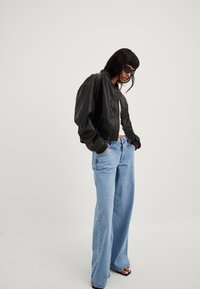 Czarna kurtka bomberka o gładkiej fakturze, w zestawieniu z jasnoniebieskimi spodniami o szerokich nogawkach. Stylizacja obejmuje czarne okulary przeciwsłoneczne i białą koszulkę.