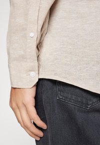 Camicia in lino beige con polsini abbottonati, che mostra una finitura in tessuto testurizzato. Mano posizionata vicino all'orlo della camicia, abbinata a jeans scuri.