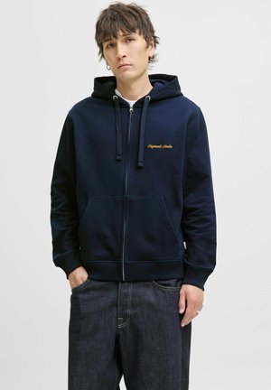 Navy zip-up hoodie in soft cotton blend, featuring drawstrings, front pockets, and embroidered text. Paired with dark denim pants.