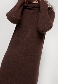 Lungo maglione marrone a maglia con collo a cowl, texture a coste e maniche lunghe. Il tessuto appare morbido e leggermente peloso.
