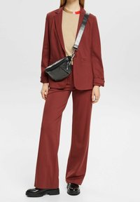 Traje ajustado burdeos con pantalones de pierna ancha y americana entallada. Top beige debajo, combinado con un pequeño bolso cruzado negro y zapatos negros gruesos.