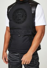 Veste matelassée noire avec accents en similicuir, logo embossé sur la poitrine et texte « GLORIOUS ». Comprend un col montant et des côtés zippés.
