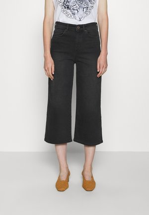 Mujer vestida con pantalones anchos de tiro corto negros, camiseta blanca con gráfico de leopardo y tacones cuadrados de yute mostaza, de pie sobre un suelo liso.