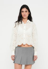 Blouse blanche courte avec broderie anglaise complexe, ourlet festonné, fermeture par boutons et manches longues, associée à une jupe plissée grise.