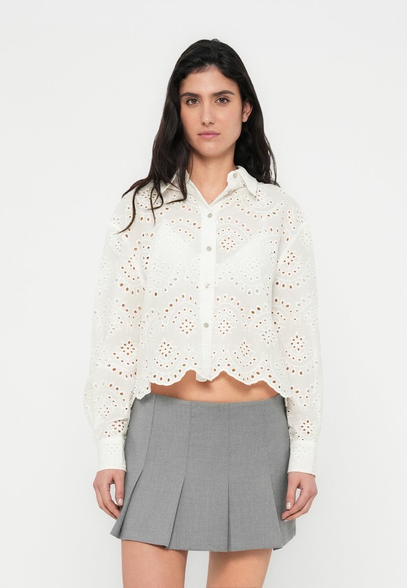 Blouse blanche courte avec broderie anglaise complexe, ourlet festonné, fermeture par boutons et manches longues, associée à une jupe plissée grise.