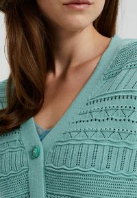Světle modro-zelený pletený cardigan s knoflíkovým zapínáním, texturovanými vzory a otevřenými stehy podél výstřihu a rukávů.