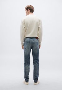 Világos bézs bordázott pulóver és fakó kék farmernadrág. A farmer szűk szabású, a hátulján barna bőr címke található. Semleges tónusú sportcipők.