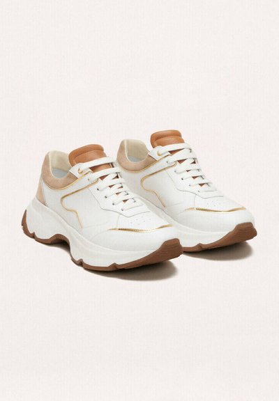 Zapatillas blancas con suela gruesa, con acentos marrones y una superficie texturada. Un ribete dorado a lo largo de los lados y detalles perforados en la puntera.