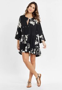 Schwarzes Blumenmuster-Kleid mit Glockenärmeln, ausgestautem V-Ausschnitt und stufigem Design. Hergestellt aus leichtem Stoff mit weißen Blumenmustern.