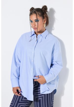 Frau mit geflochtenen Haar-Dutts, die goldene Creolen, leuchtend blaues Augen-Make-up, ein übergroßes hellblaues Hemd und gemusterte dunkle Hose trägt.