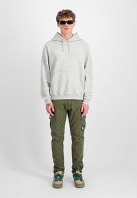Sivi hoodie sa kenguruvim džepom, maslinaste cargo hlače s aplikacijom i tenisice s zelenim akcentima. Model nosi uske sunčane naočale.