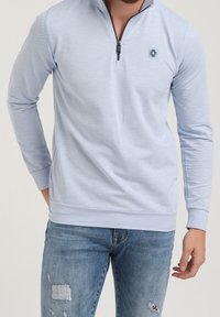 Sudadera de cremallera azul claro, hecha de tejido suave, con mangas largas, puños acanalados y un pequeño detalle de logo en el pecho. Combinada con jeans desgastados.