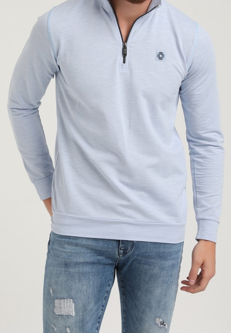 Sudadera de cremallera azul claro, hecha de tejido suave, con mangas largas, puños acanalados y un pequeño detalle de logo en el pecho. Combinada con jeans desgastados.