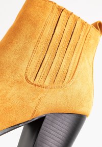 Bottine en daim orange vif avec un détail de couture verticale sur le côté, dotée d'un talon noir texturé.