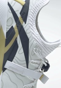 Gros plan du côté d'une chaussure de sport blanche avec un design en « X » noir et doré, une sangle élastique blanche et un verrou de lacet réglable.