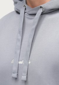 Helehall mikrotekstuuriga kapuutsiga dressipluus, millel on hõbedase metallotsikuga nöör ja valge tikandiga tekst rinnal.