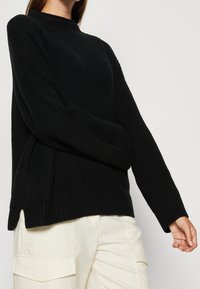 Schwarzer gerippter Strickpullover mit hohem Kragen, langen Ärmeln und seitlichen Schlitzen am Saum. Getragen mit hellen Hosen.