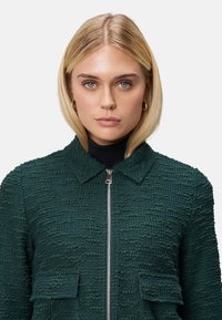 Veste verte texturée avec fermeture éclair à l'avant, col et deux poches poitrine. Présente un motif de tissu tissé et une coupe structurée.