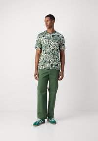 Short-sleeve green floral shirt with cream print, paired with green pants and green sneakers featuring white stripes and brown soles.