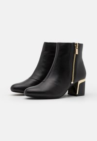 Par de botas de cuero negro hasta el tobillo con tacones de bloque y cremalleras laterales doradas sobre un fondo blanco.
