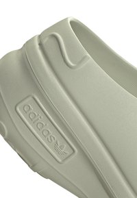 Ljusgrå slip-on sko gjord av texturerad gummi, med en upphöjd "adidas"-logotyp och en slät, rundad bakdel.