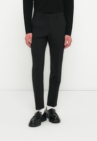 Fekete slim-fit nadrágot, fekete mokaszin cipőt, fehér zoknit és fekete hosszú ujjú felsőt viselő személy áll egy sima fehér háttér előtt.