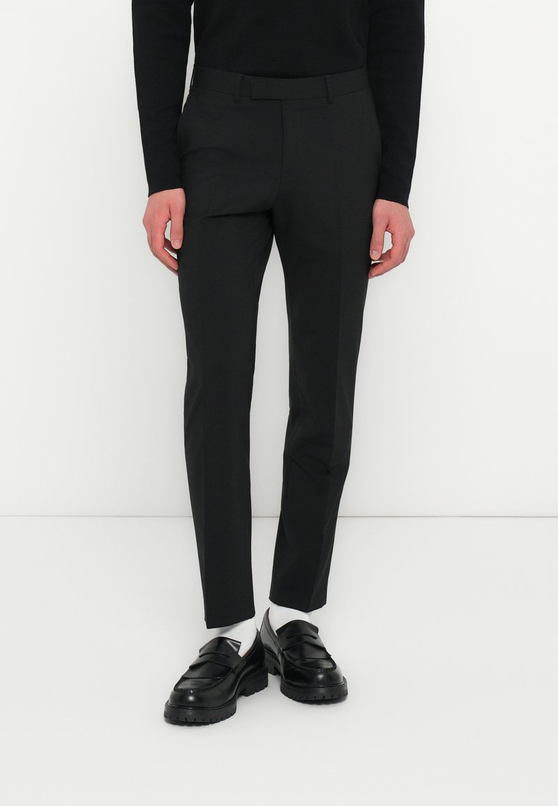 Fekete slim-fit nadrágot, fekete mokaszin cipőt, fehér zoknit és fekete hosszú ujjú felsőt viselő személy áll egy sima fehér háttér előtt.