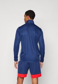 Marineblå atletisk langermet topp med høyt krage, glatt tekstur, kontrasterende hvite sidestriper, parret med marineblå shorts med røde detaljer.
