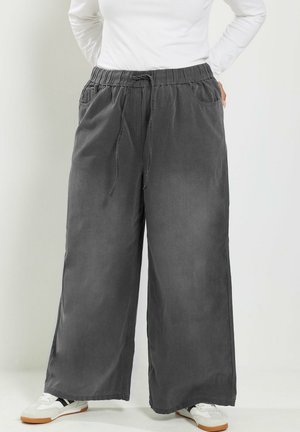 Person trägt weit geschnittene dunkelgraue Hose mit Kordelzug in der Taille, weißes Langarmhemd und weiße Sneakers mit schwarzen Streifen.