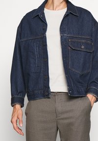 Ciemnoniebieska kurtka dżinsowa z kołnierzem, zapinana na guziki z przodu, z kieszenią na piersi i kontrastowymi żółtymi szwami, noszona na jasnym t-shircie.