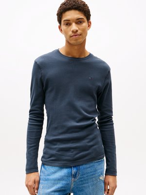 Ung man klädd i en marinblå långärmad skjorta och ljusblå jeans, stående framför en enfärgad vit bakgrund.