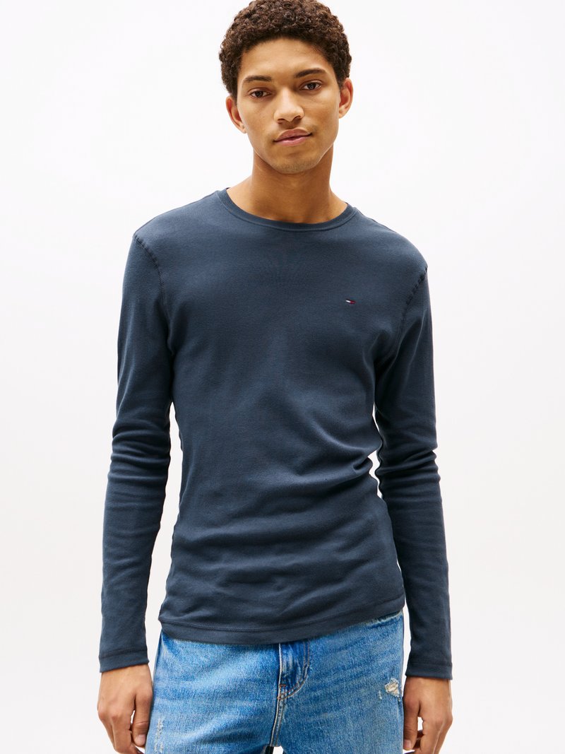 Ung mann iført en marineblå langermet skjorte og lyseblå jeans som står mot en ensfarget hvit bakgrunn.