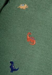 Strickstoff in Grün mit bunten, gestickten Dinosauriermotiven in Orange, Blau und Beige, die ausgeprägte Formen und Muster aufweisen.