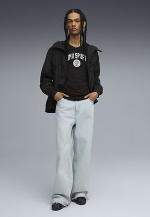 Sort letvægtsjakke over en sort t-shirt med hvidt logo, parret med lyseblå wide-leg jeans og sorte sko med blå accenter.